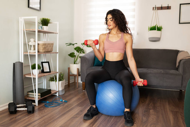 Gut aussehende hispanische junge Frau, die in ihrem heimischen Wohnzimmer in einem Fitnessball sitzt. Aktive Frau in ihren Zwanzigern beim Training mit Hantelgewichten - Foto, Bild
