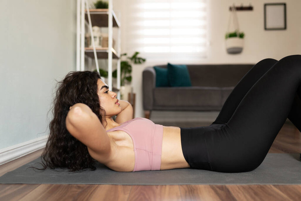 Вид сбоку на стройную молодую женщину, сидящую на коврике для упражнений в своем доме - Фото, изображение