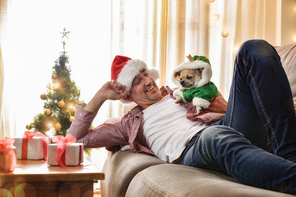 Мужчина лежит дома на диване со своей собакой, одетой в рождественский костюм с украшенной елкой и подарками. - Фото, изображение