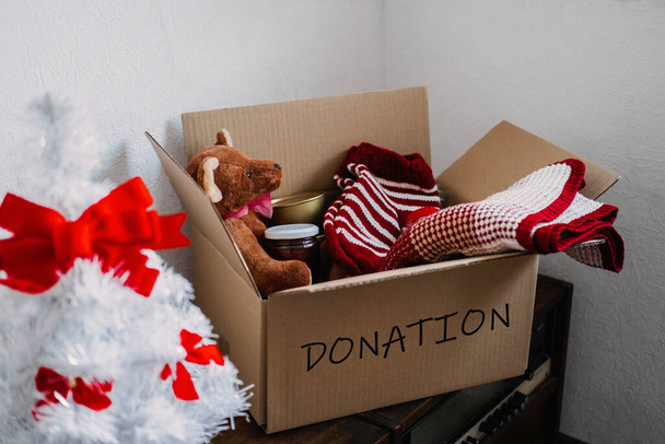 Рождественские корзины, помощь беженцам и бездомным. Рождественская коробка для пожертвований с теплой одеждой, едой и игрушками у елки дома. Благотворительные организации, дающие рождественские корзины нуждающимся семьям - Фото, изображение