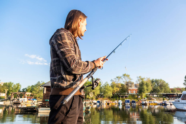 Mid age white man with long brown hair is setting fishing rod at river bank in spring. Boats and floating houses are around him with trees in background. He is using fishing rod with spinning reel, green line and lure. - Photo, Image