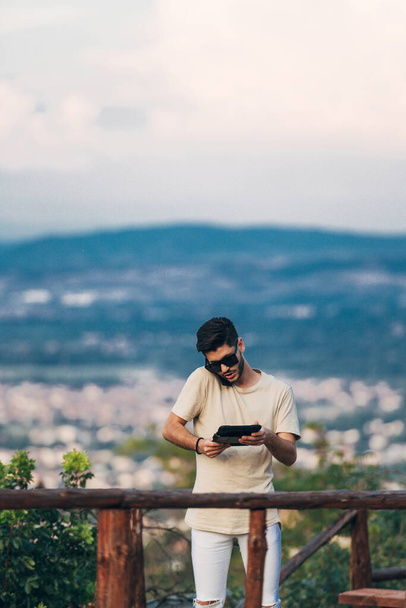 Hombre sentado en una mesa de madera usando una tableta digital mientras mantiene una conversación móvil en la colina - Foto, Imagen