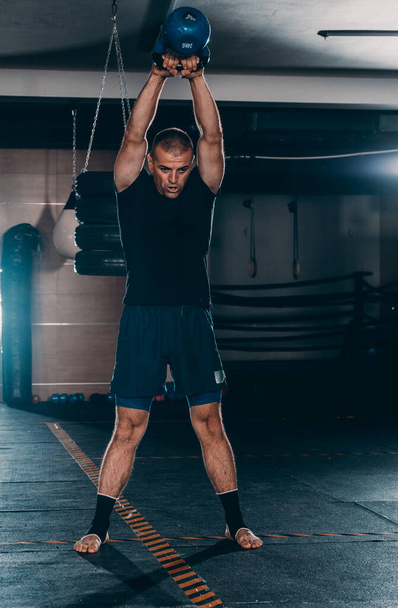 Handsome athletic man holding kettlebell overhead while working out in dark gym - Photo, Image