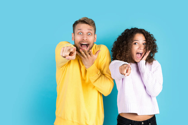 Choose new advertising options. Excited and shocked man together with happy african american woman, pointing at camera, making amazed face, standing on blue background with copy space - Fotó, kép