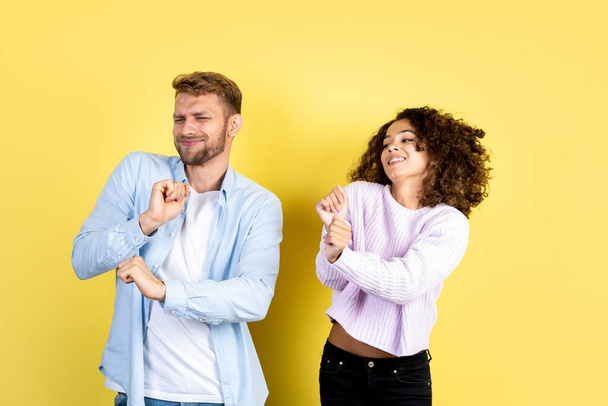 Two stylish mixed race friends listening music, moving together, dancing on party, standing on yellow background with copy space. Happy people celebrating winning a prize or birthday - Photo, Image