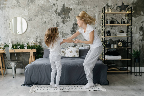 Glückliche Kindheit. Ganztägige Ansicht der fröhlichen Mutter, die mit ihrer kleinen Tochter tanzt, neben dem Bett springt, Spaß hat und den Morgen zu Hause zusammen verbringt - Foto, Bild