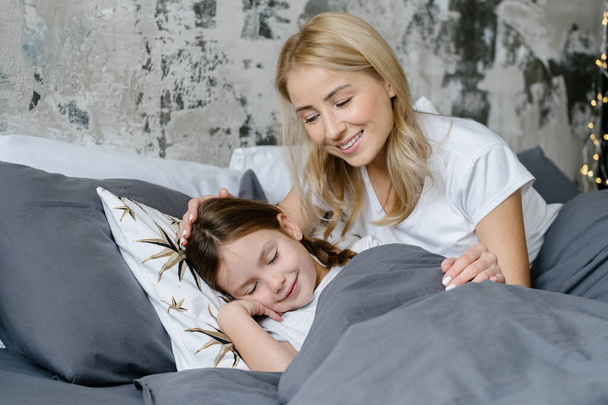 Portret samotnej matki głaskającej uśmiechniętą córkę po głowie, spędzającej razem poranek w przytulnym domu podczas snu dziecka. Kobieta z dzieckiem leży w wygodnym łóżku - Zdjęcie, obraz
