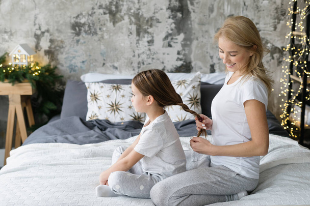 Side view of caring mother braids hair and making hairstyle to her smiling daughter. Girl and mom sitting on bed in pajamas, spending morning in bedroom together, resting at home - Photo, Image