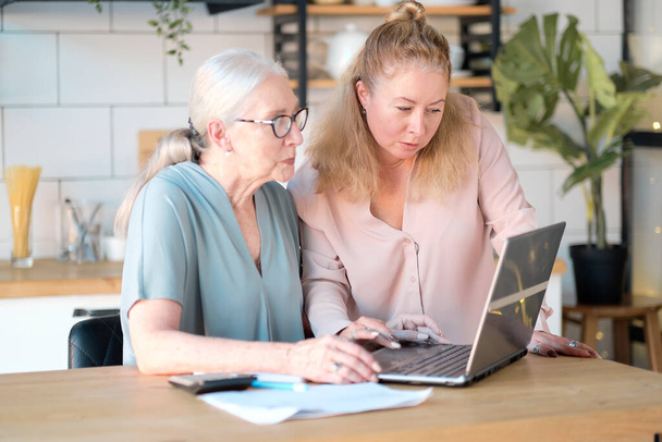 daughter helps her elderly mother figure it out online with her personal account. Woman teaching senior mother to use internet at home. Senior woman with her daughter looking at modern gadget indoors. - Photo, Image