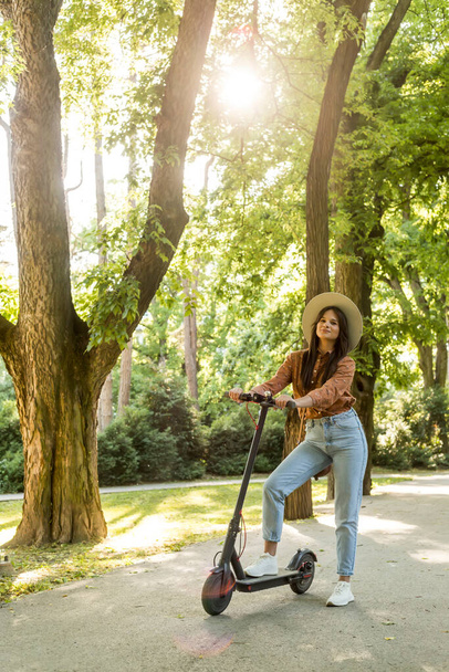 A young beautiful woman with a hat spending sunny spring day in the park and posing on her electric scooter, while trees predominate in the background. - Foto, imagen
