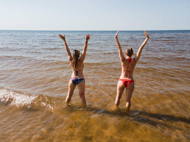 Vista aérea trasera: Dos mujeres en trajes de baño se paran y levantan las manos en agua de mar - Foto, Imagen