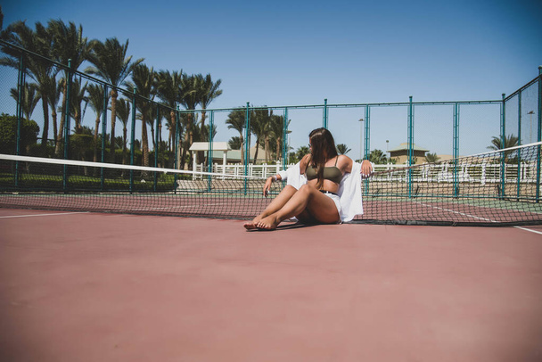 Stylish cute teenage model dressed in summer hipster vintage white stylish clothes. Trendy girl posing in sunglasses sitting on a tennis court. The original shadow from the grid at the sports field - 写真・画像