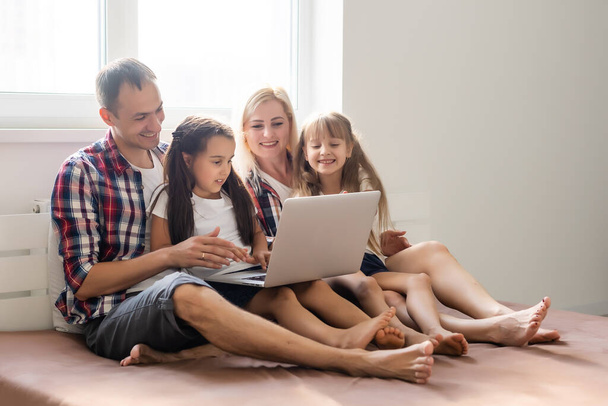 Glückliches Familienkonzept. Schöne Mutter und schöner Vater mit ihren Töchtern, die Zeit zu Hause verbringen und mit Laptop im Bett liegen. - Foto, Bild