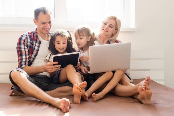 Boldog családi koncepció. Gyönyörű anya és jóképű apa a lányaikkal együtt töltik az időt otthon és az ágyon fekszenek laptoppal.. - Fotó, kép