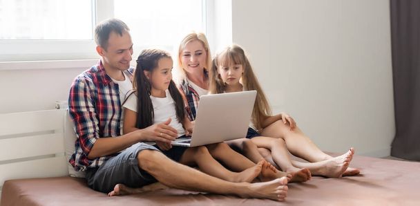 Happy family concept. Beautiful mother and handsome father with their daughters spending time together at home and lying on bed with laptop. - Fotoğraf, Görsel