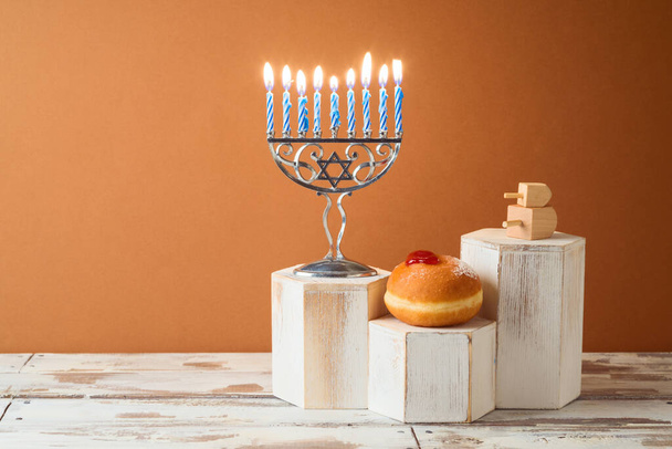Jewish holiday Hanukkah creative concept with menorah and traditional donuts on podium on wooden table - Zdjęcie, obraz