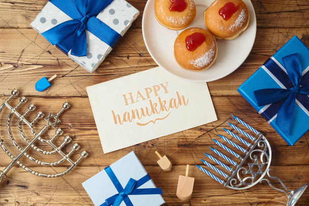 Jewish holiday Hanukkah greeting card with traditional donuts, menorah and gift box on wooden background - 写真・画像