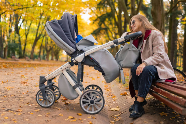 Cheerful mother sits on bench in park and looks on the baby who sleeps in stroller - Photo, Image