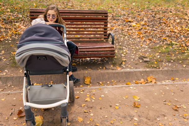 Bebek arabalı anne parkta bankta oturur ve bebek arabasında uyuyan bebeğe bakar. - Fotoğraf, Görsel