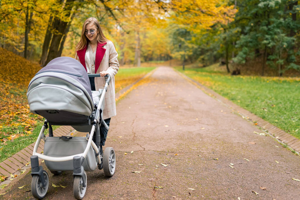 Cheerful mother with stroller walks in  autumn park away from the bustle of the city - Фото, изображение