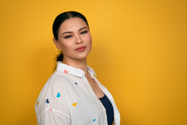 Charming Asian woman standing sideways looking at camera, wearing casual white shirt, isolated on yellow background. Human emotions, facial expression concept - Photo, image