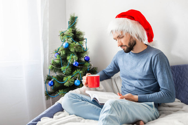 Junger kaukasischer Mann sitzt auf Sofa, liest Buch und hält Tasse Kaffee zu Hause auf Weihnachtsbaum Hintergrund. - Foto, Bild
