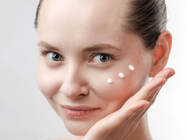 Beautiful Young Woman with Clean Fresh Skin. Drops of cream are applied on the cheek. Hand near the face. Girl beauty face care. Facial treatment . Cosmetology , beauty and spa. Natural cosmetic product. Concept of Organic, Bio Cosmetics. Isolated on - Foto, Bild