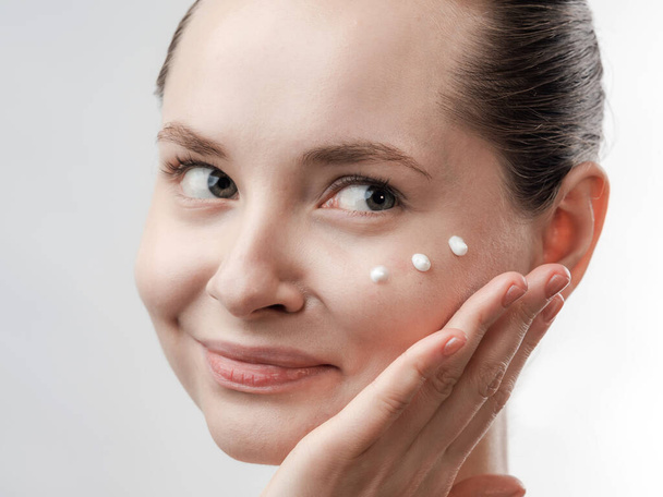Beautiful Young Woman with Clean Fresh Skin. Drops of cream are applied on the cheek. Hand near the face. Girl beauty face care. Facial treatment . Cosmetology , beauty and spa. Natural cosmetic product. Concept of Organic, Bio Cosmetics. Isolated on - Foto, immagini