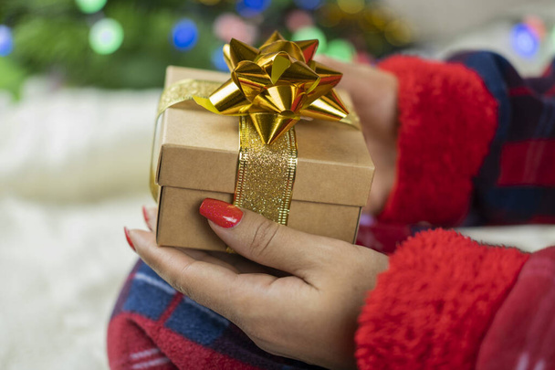 девушка в пижаме Санта-Клауса с коробкой сюрприза с золотым бантом на новогодний праздник на фоне новогодней елки с гирляндой - Фото, изображение