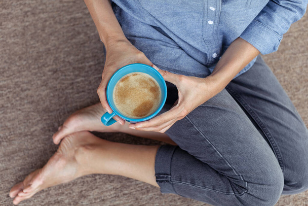 Női kéz, kezében egy csésze kávéval. Egy fiatal nő ül a földön. A lány, aki kávét iszik vagy teát, otthon ül. Nyugodj meg és pihenj. Közelkép. - Fotó, kép