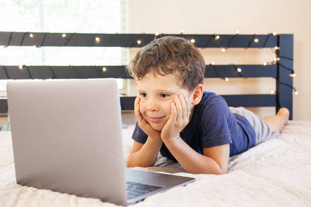 Uroczy chłopiec leżący na przodzie w łóżku i patrzący na ekran komputera. Edukacja domowa i koncepcja wolnego czasu z komputerem u dzieci - Zdjęcie, obraz