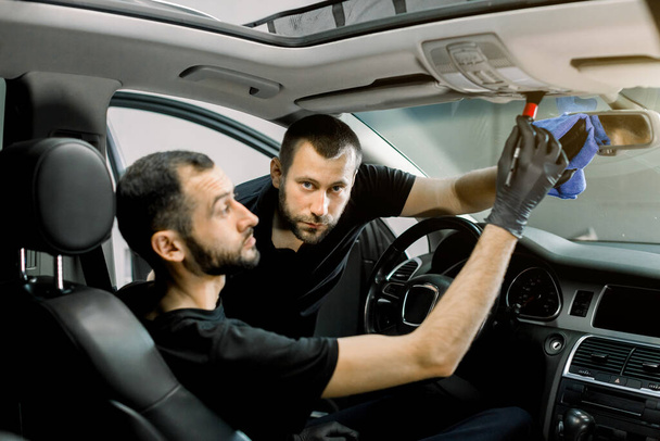 Two handsome male auto service workers cleaning modern car interior. Focus on the man, cleaning car mirror with microfiber cloth, while his colleague removes the dust from roof plastic panel - Fotografie, Obrázek