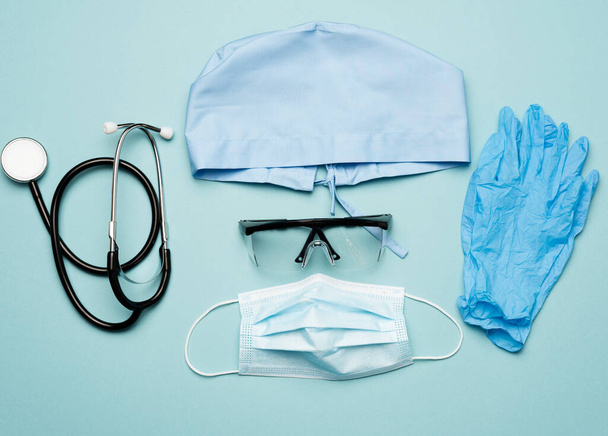υφασμάτινο μπλε καπέλο, ιατρική μάσκα μιας χρήσης, ζευγάρι γάντια και πλαστικά γυαλιά σε μπλε φόντο, πάνω όψη. Προστατευτική ενδυμασία γιατρού - Φωτογραφία, εικόνα