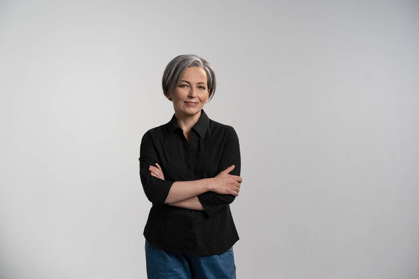Pretty mid aged grey haired woman in black shirt isolated on grey background. Confident mature grey haired woman looking on camera with arms folded. Human emotions, facial expression concept - Фото, зображення