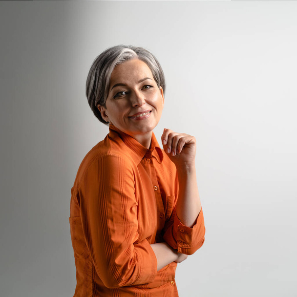 Portrait of a mature grey haired woman wearing orange shirt happily smiling on camera. Pretty mid aged grey haired woman in orange shirt isolated on grey background - Φωτογραφία, εικόνα