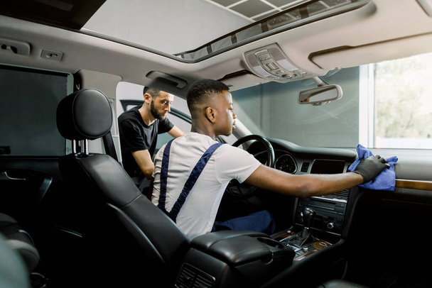 Car service and detailing station. Back angle indoor shot of two professional multiethnic men workers, providing a vehicle interior cleaning, wiping front panel with microfiber cloth and brush - Foto, Bild