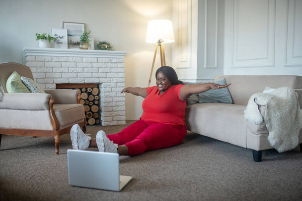 Plus size african american woman having an online workout and stretching her arms to the sides - Photo, image