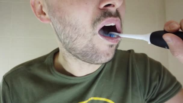 Een man met een baard poetst zijn tanden in de spiegel met zijn mond open. Tandpasta op een elektrische tandenborstel - Video