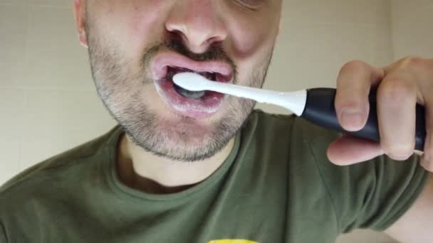 Un uomo barbuto si lava i denti nello specchio con la bocca aperta. Dentifricio su uno spazzolino elettrico - Filmati, video