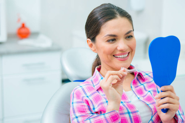 Positive woman sitting in a dental chair with a dental plate in hand and looking at her reflection in the mirror - Foto, Imagen