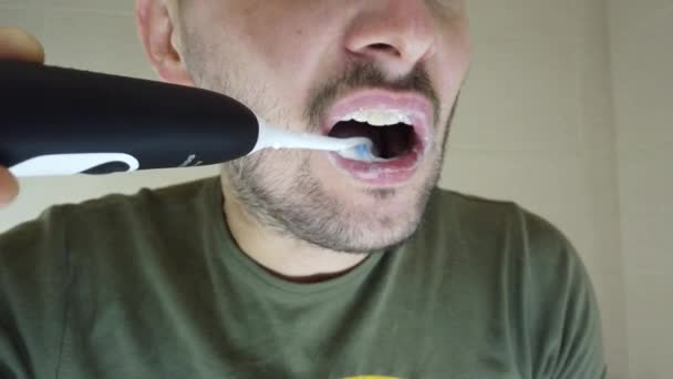 Un barbudo se cepilla los dientes en el espejo con la boca abierta. Pasta de dientes en un cepillo de dientes eléctrico - Imágenes, Vídeo