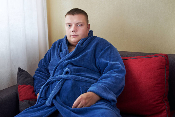 Толстяк в халате сидит дома на диване, неправильный образ жизни. - Фото, изображение