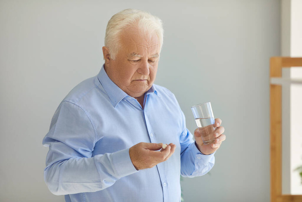 Hombre mayor sosteniendo píldora médica y vaso de agua dudando si el medicamento vale la pena - Foto, Imagen