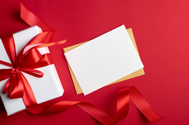 Greeting card mockup. White gift box with red ribbon and bow on red paper background. Christmas and New Year holidays concept, top view, copy space. - Foto, Bild