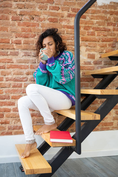 Jovem bebendo café. Ela está sentada nas escadas de sua casa ao lado de um livro. - Foto, Imagem