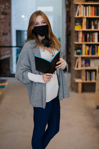 Молодая блондинка в маске с книгой в книжном магазине, смотрящая в камеру. - Фото, изображение