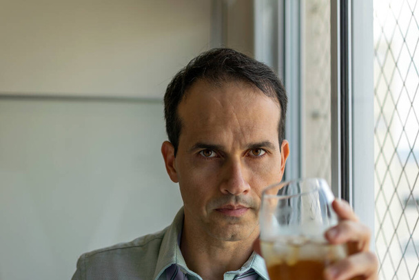 Κοντινό πλάνο ενός ώριμου άνδρα (44 ετών) που κρατάει ένα ποτήρι ουίσκι δίπλα σε ένα παράθυρο. - Φωτογραφία, εικόνα
