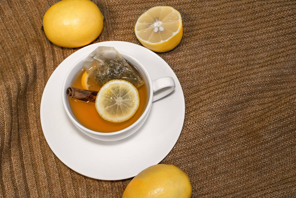 Lemon tea with cinnamon in a white Cup on a saucer stands on a warm sweater with lemons - Photo, Image