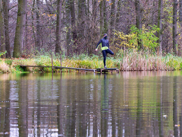 湖のモルや桟橋の外にストレッチ黒い緑のジャケットでアスリート。霧の秋の自然を走るクロスカントリーのためのトレイルランナーのトレーニング. - 写真・画像