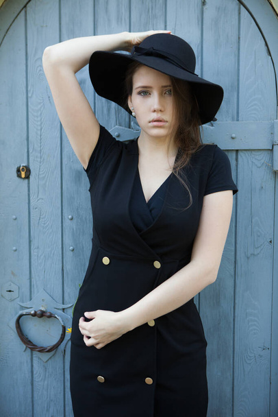 Beautiful young woman in a black dress and a black hat against the old door - Фото, зображення